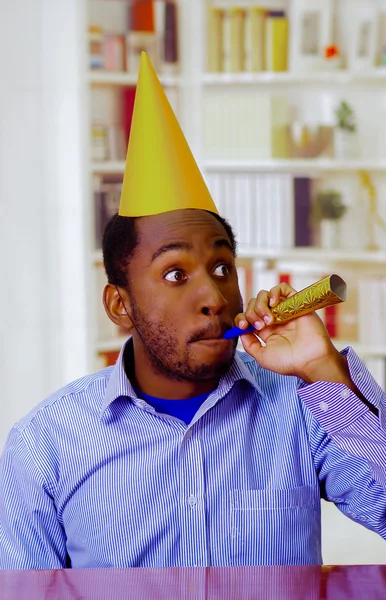 Homem encantador vestindo camisa azul e chapéu sentado à mesa soprando chifre festa celebrando sozinho sorrindo — Fotografia de Stock