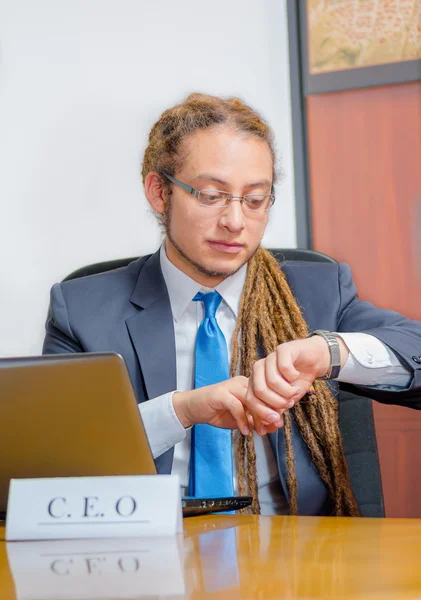 Handsome man with dreads and business suit sitting by desk looking at wrist watch, young manager concept — Stock Photo, Image