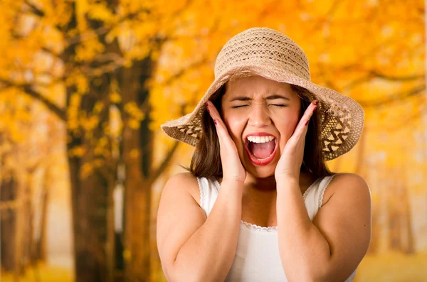 Young attractive woman wearing white top and fashionable hat, touching cheeks with own hands screaming out, forest autumn background — Stock Photo, Image