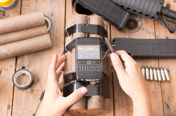 Руки с самодельной бомбой с взрывчаткой и сотовым телефоном прикреплены провода сидя на поверхности рядом с другими используемыми частями, пулемет на заднем плане — стоковое фото