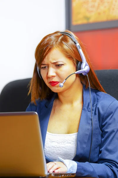 Junge attraktive Frau in Bürokleidung und Headset sitzt am Schreibtisch und schaut auf den Computerbildschirm, arbeitet mit frustriertem Gesichtsausdruck — Stockfoto