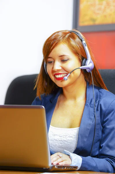 Young attractive woman wearing office clothes and headset sitting by desk looking at computer screen, working with positive facial expression