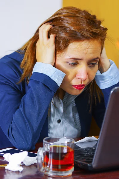 Mulher jovem doente de gripe, sentada à mesa usando o computador trabalhando, olhando mal e cansado, medicamentos, tecidos, vidro de água na mesa — Fotografia de Stock