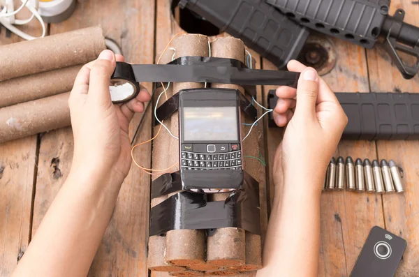 Hands handling homemade bomb with explosives and cellular phone attached wires sitting on surface next to other used parts, machine gun in background — Stock Photo, Image