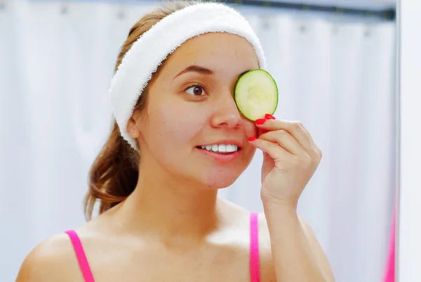 Mulher jovem atraente vestindo top rosa e cabeça branca, cobrindo um olho com fatia de pepino usado para o tratamento da pele, olhando no espelho sorrindo — Fotografia de Stock