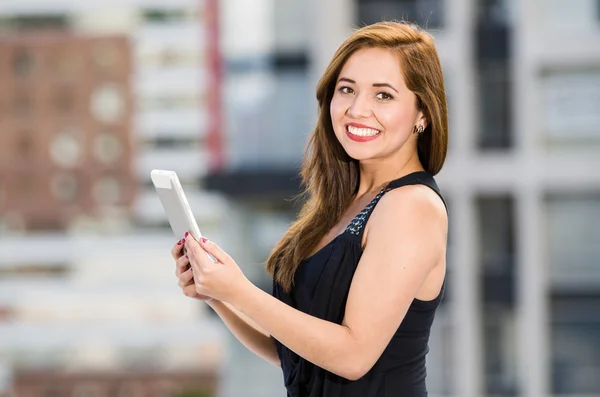 Jovem mulher atraente vestindo vestido preto em pé no telhado, segurando tablet, sorrindo para a câmera, edifícios da cidade fundo — Fotografia de Stock