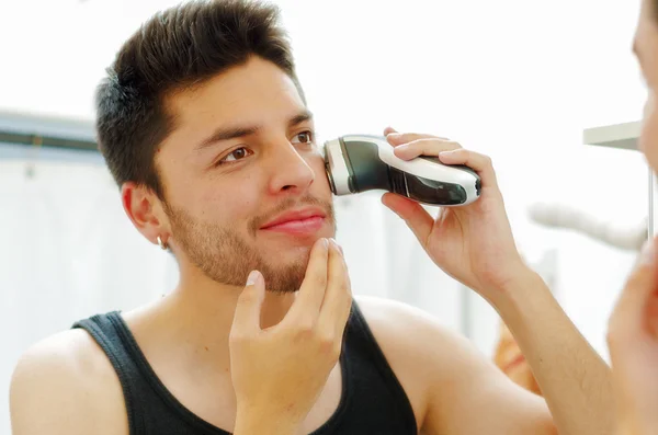 Przystojny młody człowiek ubrany w czarny singletowego Top patrząc w lustro, za pomocą elektrycznych Maszynka do golenia podczas porannej koncepcji rutynowych, lekko uśmiechnięty — Zdjęcie stockowe