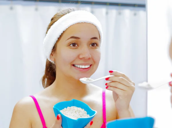 Привлекательная молодая женщина в розовой верхней и белой повязке, держит ложку с йогуртом, глядя в зеркало улыбаясь — стоковое фото