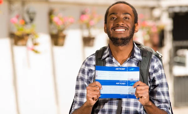 Joven hombre feliz vistiendo ropa casual y mochila, mostrando boletos de viaje para la cámara mientras sonríe, ambiente de jardín, concepto de mochilero — Foto de Stock