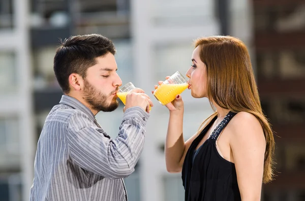 Młoda atrakcyjna para ubrana w formalne ubrania stojąca na dachu twarzą do siebie pijąca ze szkła z żółtym napojem, pozująca do kamery, tło budynków miejskich — Zdjęcie stockowe