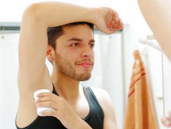 Yakışıklı genç adam siyah singlet üst aynaya bakarak giyiyor, sabah rutin kavramı sırasında deodorant uygulayarak, sligthly gülümseyerek — Stok fotoğraf