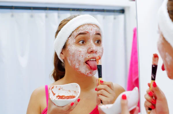 Atractiva mujer joven con top rosa y diadema blanca usando cepillo para aplicar crema en la cara, sosteniendo la lengua simulando comer, mirando en el espejo — Foto de Stock