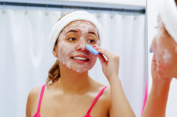 Attraktive junge Frau mit rosa Oberteil und weißem Stirnband, die mit einem Schwamm Creme aus dem Gesicht entfernt und lächelnd in den Spiegel schaut — Stockfoto