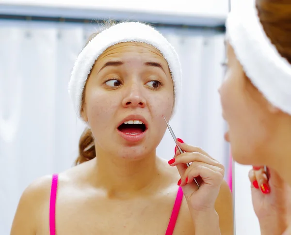 Wanita muda yang menarik mengenakan ikat kepala merah muda dan putih, menyentuh wajah dengan alat perawatan kulit, melihat di cermin berkonsentrasi — Stok Foto
