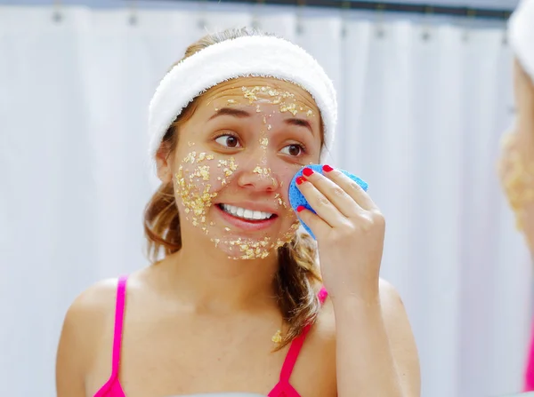 Attractive young woman wearing pink top and white headband, removing oat mixture from face using sponge, looking in mirror smiling — Stock Photo, Image