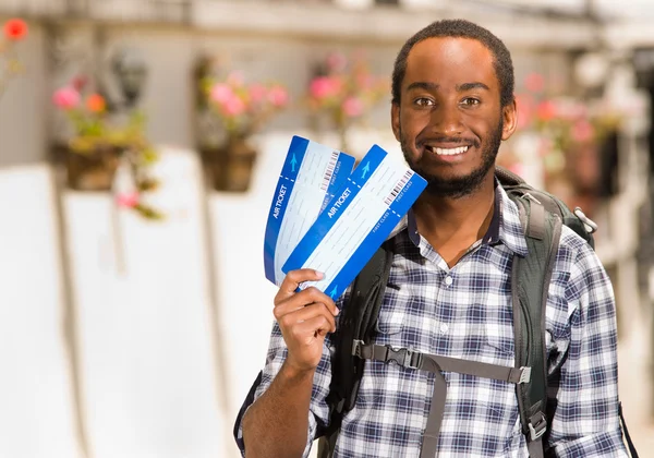 Jeune homme heureux portant des vêtements décontractés et sac à dos, montrant les billets de voyage pour la caméra tout en souriant, environnement de jardin, concept de routard — Photo