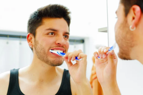 Handsome young man wearing black singlet top looking in mirror smiling while brushing teeth