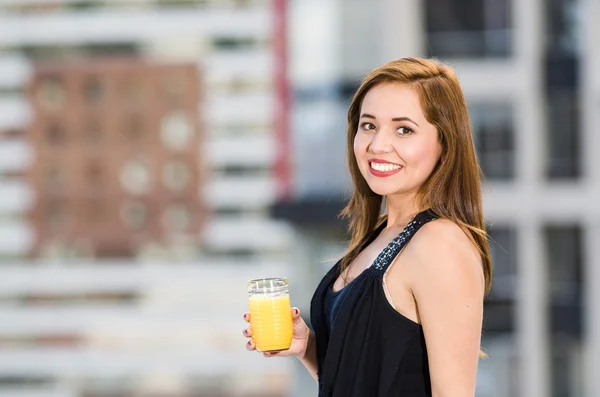 Mladá atraktivní žena v černých šatech stojící na střeše, drží sklo se žlutým nápojem, usmívá se do kamery, městské budovy pozadí — Stock fotografie