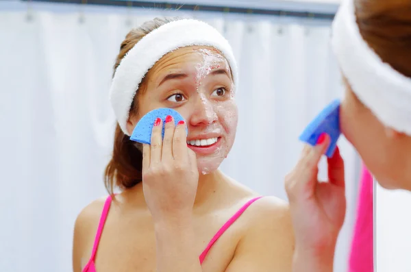 Attractive young woman wearing pink top and white headband removing cream from face using sponge, looking in mirror smiling — Stock Photo, Image