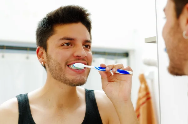 Bonito jovem vestindo top singlet preto olhando no espelho sorrindo enquanto escova os dentes — Fotografia de Stock