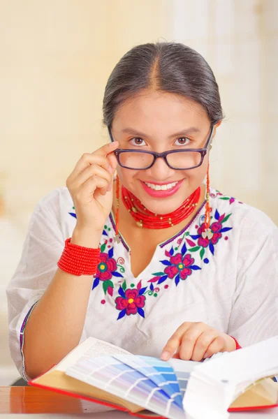 Young pretty girl wearing white shirt with colorful flower decorations and glasses, sitting by desk reading book smiling, bright background — Stock Photo, Image