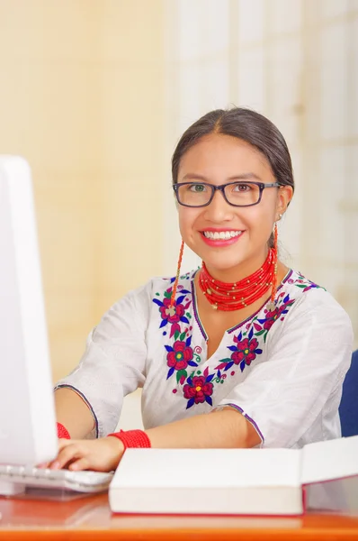 Młoda ładna dziewczyna w białej koszuli z kolorowe dekoracje kwiatowe i okulary, siedzi przy biurku wpisując na klawiaturze komputera uśmiechnięty, otwarta książka przed — Zdjęcie stockowe