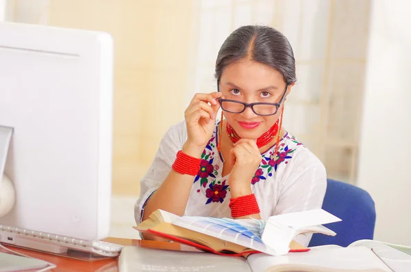 Young pretty girl wearing white shirt with colorful flower decorations and glasses, sitting by desk reading book smiling, bright background — Stock Photo, Image