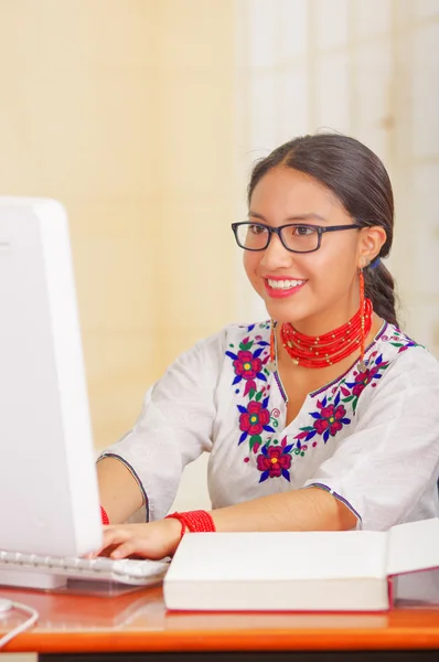 Giovane bella ragazza indossa camicia bianca con decorazioni floreali colorate e occhiali, seduto alla scrivania digitando sulla tastiera del computer sorridente, libro aperto di fronte — Foto Stock