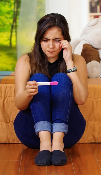 Mulher morena muito jovem sentada no chão segurando teste de gravidez em casa, chorando enxugando lágrimas, olhando emocional, fundo da janela do jardim — Fotografia de Stock
