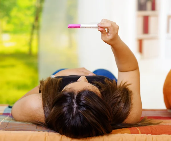 Jolie jeune femme brune allongée tenant le test de grossesse à la maison devant, vue de derrière la tête, les étagères et le fond du jardin de la fenêtre — Photo