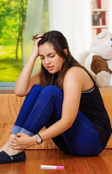 Hübsche junge brünette Frau auf dem Boden sitzend mit Schwangerschaftstest vor sich liegend, schockiert verzweifelt blickend, Gartenfenster-Hintergrund — Stockfoto