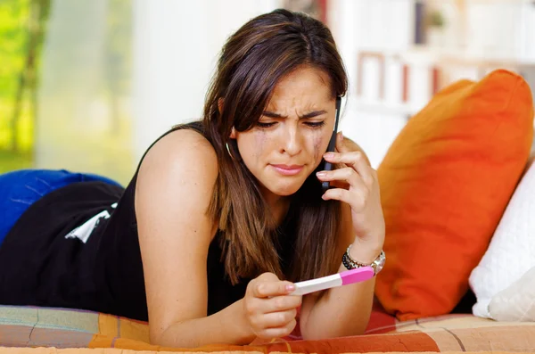 Jolie jeune femme brune allongée tenant un test de grossesse à la maison devant, parlant sur mobile, étagères et fond de jardin fenêtre — Photo