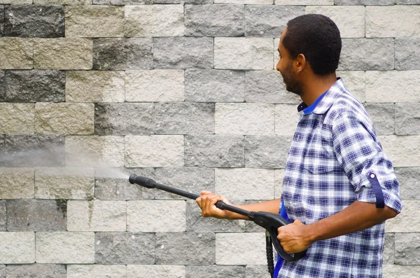 Hombre con patrón cuadrado camisa azul y blanca sosteniendo pistola de agua de alta presión, apuntando hacia la pared de ladrillo gris — Foto de Stock