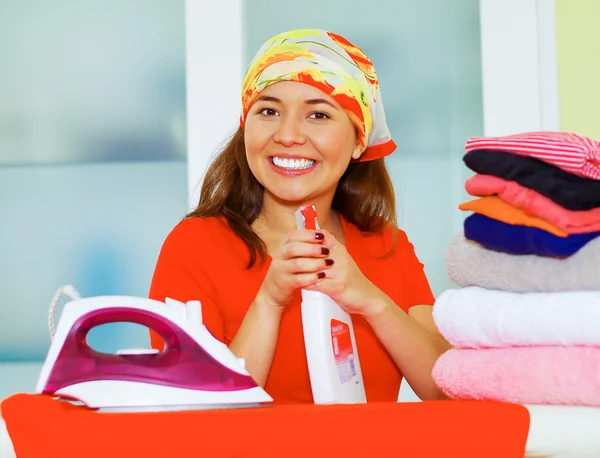 Joven encantadora mujer con colorido pañuelo para la cabeza planchado pila de ropa mientras sonríe felizmente, lavandería concepto de tareas domésticas — Foto de Stock