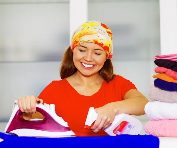 Joven encantadora mujer con colorido pañuelo para la cabeza planchado pila de ropa mientras sonríe felizmente, lavandería concepto de tareas domésticas — Foto de Stock