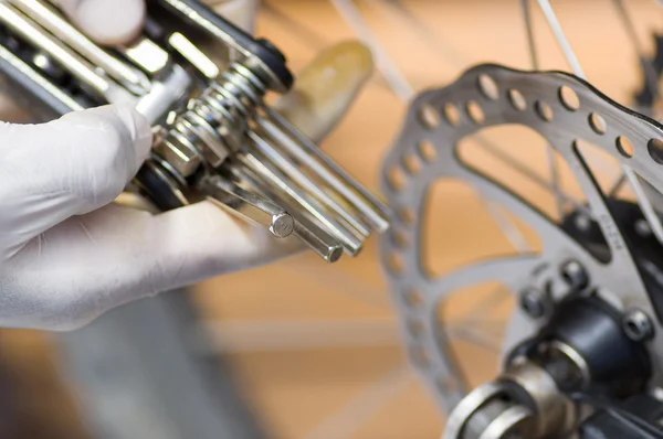 Mão close-up vestindo luva branca segurando multi unbrako ferramenta chave ao lado de partes de roda de bicicleta, conceito de reparo mecânico — Fotografia de Stock