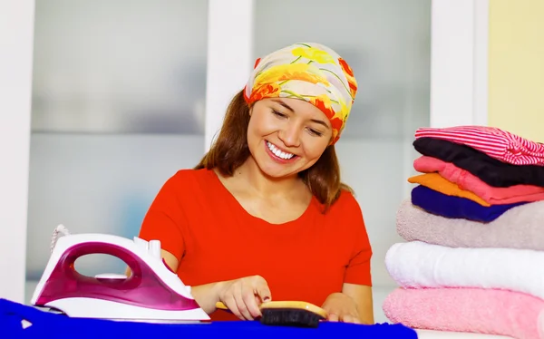 Joven encantadora mujer con colorido pañuelo para la cabeza planchado pila de ropa mientras sonríe felizmente, lavandería concepto de tareas domésticas — Foto de Stock