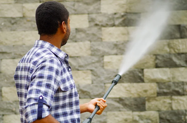 Hombre con patrón cuadrado camisa azul y blanca sosteniendo pistola de agua de alta presión, apuntando hacia la pared de ladrillo gris — Foto de Stock