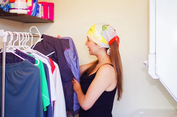 Encantadora mujer joven con colorido pañuelo para la cabeza de pie dentro del armario mirando a través de la selección de ropa og colgando, concepto de tareas domésticas de lavandería — Foto de Stock