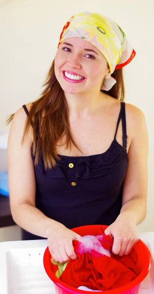 Encantadora joven con pañuelo colorido en la cabeza sonriendo a la cámara mientras se lava las manos algo de ropa, concepto de tareas domésticas de lavandería — Foto de Stock