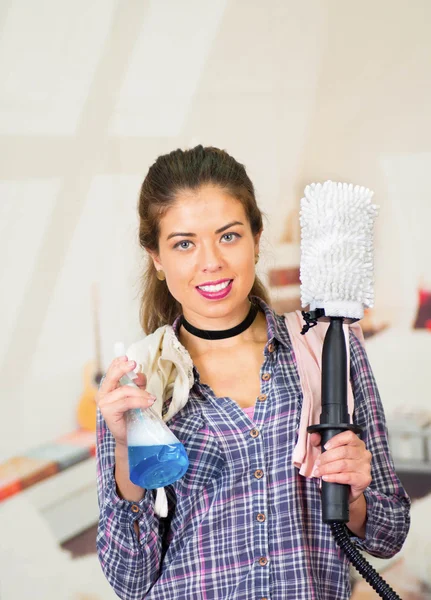 Atractiva morena vestida con ropa casual sosteniendo la cabeza del dispositivo de lavado eléctrico con manguera conectada, sonriendo y posando para la cámara, fondo de apartamento desordenado —  Fotos de Stock