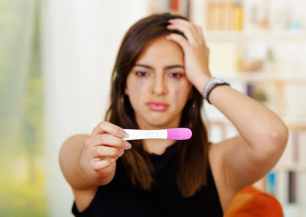 Pretty young brunette woman holding pregnancy home test, staring at it crying wiping tears. looking emotional, garden window background — Stock Photo, Image