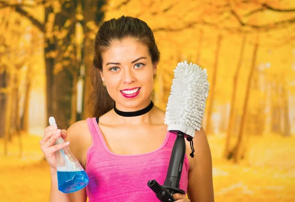 Attractive brunette wearing pink top posing while holding up cleaning product bottle and mop device which connects to water hose, autumn forest background — Stock Photo, Image