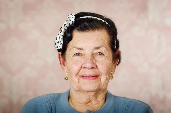 Пожилая милая латиноамериканка в голубом свитере и галстуке в горошек на голове, осторожно улыбающаяся перед розовыми обоями — стоковое фото