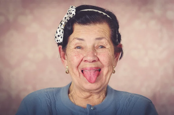 Femme hispanique mignonne plus âgée portant un pull bleu et un nœud papillon à pois sur la tête montrant sa langue à l'appareil photo devant un papier peint rose — Photo