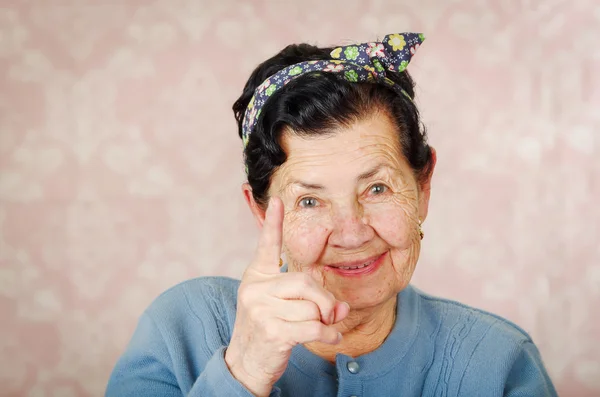 Older cute hispanic woman wearing blue sweater and flower pattern bow on head holding up one finger for the camera in front of pink wallpaper