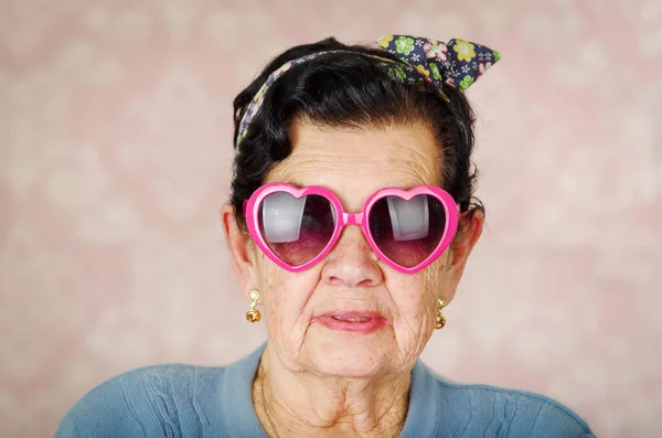 Femme hispanique cool plus âgée portant un pull bleu, un arc à motif de fleurs sur la tête et des lunettes de soleil roses en forme de cœur regardant dans la caméra — Photo