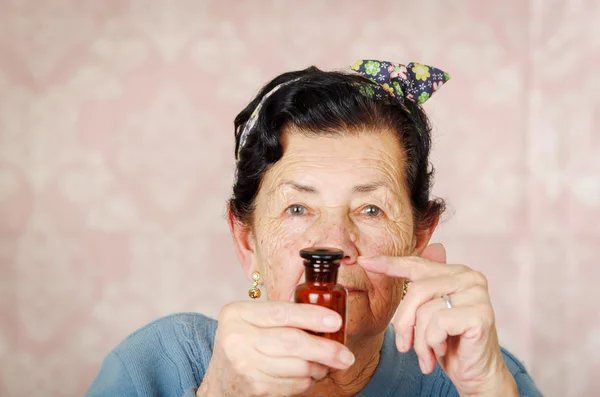 Old cool hispanik wanita mengenakan sweater biru, pola bunga busur di kepala memegang botol kaca merah kecil untuk kamera — Stok Foto