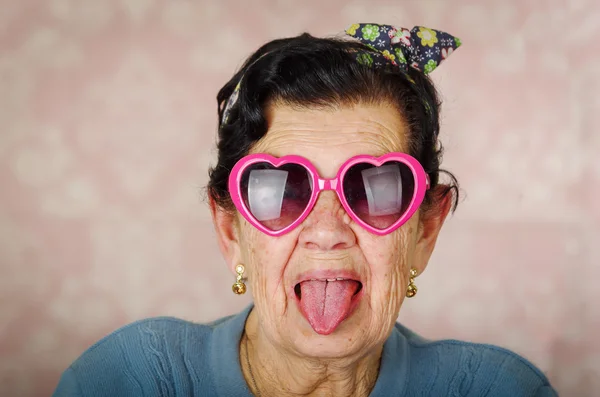 Older cool hispanic woman wearing blue sweater, flower pattern bow on head and pink heartshaped sunglasses looking into camera showing her tongue — Stock Photo, Image