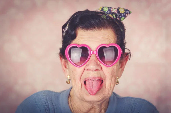Starší cool hispánský žena na sobě modrý svetr, květinový vzor bow na hlavy a sluneční brýle růžové srdíček při pohledu do kamery, zachycující její jazyk — Stock fotografie